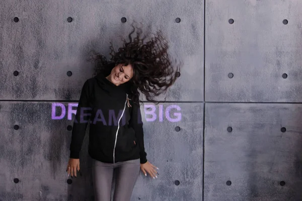 Mulher jovem encaracolado com cabelo voador posando ativamente no fundo da parede de concreto. Um sonho iluminado colorido grande . — Fotografia de Stock