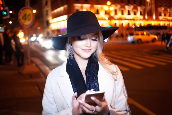 Snygg ung kvinna i en elegant beige rock använder en smartphone står vid vägen, på gatan i staden på natten. — Stockfoto