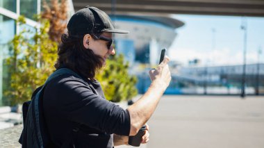 Siyah gözlüklü, siyah beysbol şapkalı ve akıllı telefonuyla sokakta kahve içen bir adam. Yan görünüm.