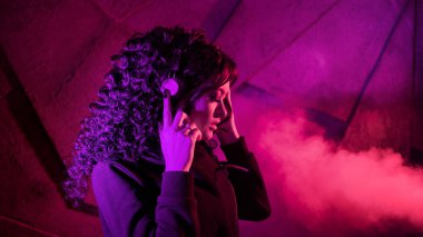 Büyük kulaklıklı genç, güzel, kıvırcık kadın müzik dinliyor, şarkı söylüyor, arka planda dans ediyor..