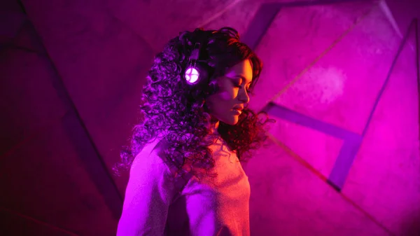 Ritratto di una donna riccia pensosa e sensuale in grandi cuffie, con un sorriso morbido ascoltando musica, su sfondo neon . — Foto Stock