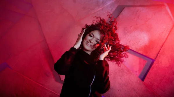 Young beautiful curly woman in big headphones smiles, listens to music, sings along, dances, jumps, closing her eyes.