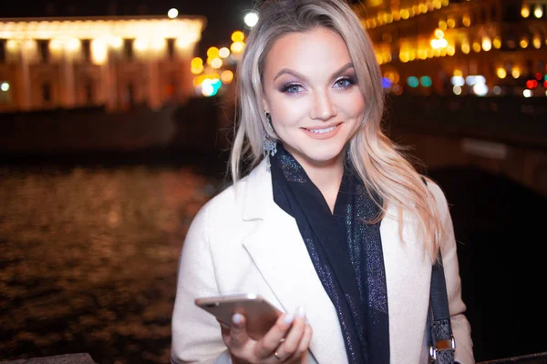Mujer joven con estilo en un elegante abrigo beige utiliza un teléfono inteligente de pie junto a la carretera, en la calle de la ciudad por la noche . — Foto de Stock