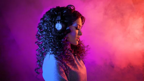 Portrait of a curly pensive sensual woman in big headphones, with a soft smile listening to music, on neon background. — Stock Photo, Image