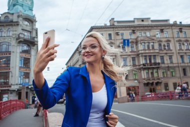 Şık gözlüklü genç kadın selfie çekiyor, tarihi binaların arka planına karşı fotoğraf çekiyor.