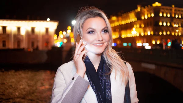 Mujer joven con estilo en un elegante abrigo beige utiliza un teléfono inteligente de pie junto a la carretera, en la calle de la ciudad por la noche . —  Fotos de Stock