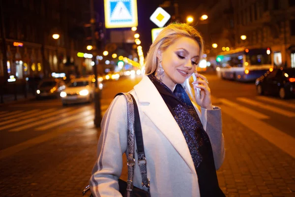 Jovem elegante em um casaco bege elegante usa um smartphone em pé na estrada, na rua da cidade à noite . — Fotografia de Stock