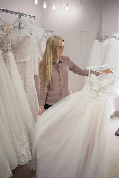 Jeune femme blonde choisit une robe de mariée dans un salon de mariage. — Photo