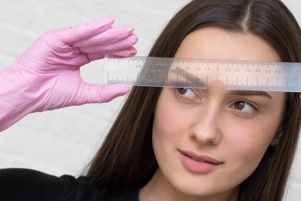 Que tipo de forma de sobrancelha está tendendo nesta temporada Uma jovem linda garota morena com uma régua de um mestre artista de sobrancelha — Fotografia de Stock