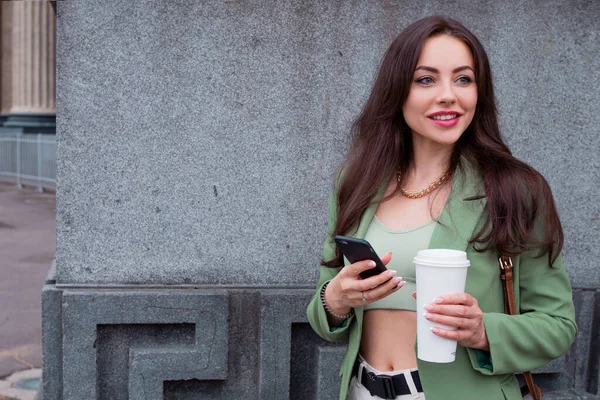 Jonge brunette vrouw in een groen jasje op een wandeling. Een meisje met een afhaalkoffie in haar handen, — Stockfoto