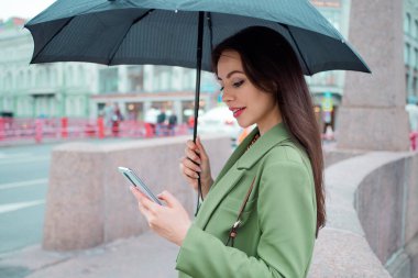 Yürüyüşe çıkmış yeşil ceketli esmer bir kadın. Şemsiyenin altındaki güzel bir kız akıllı telefon kullanır.