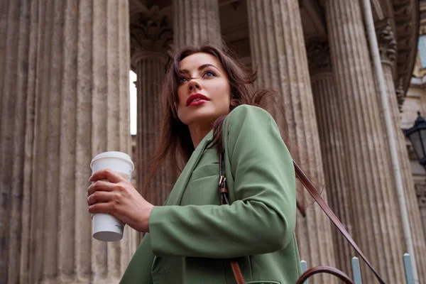Junge brünette Frau in grüner Jacke auf einem Spaziergang. Business-Mädchen mit Kaffee zum Mitnehmen, — Stockfoto