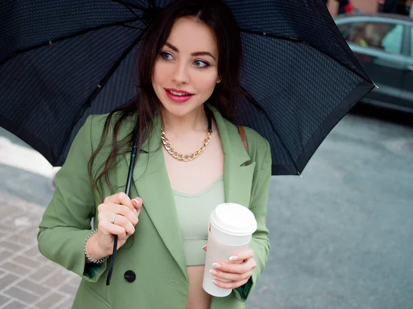 Jovem morena em uma jaqueta verde em uma caminhada. Menina bonita com café takeaway — Fotografia de Stock