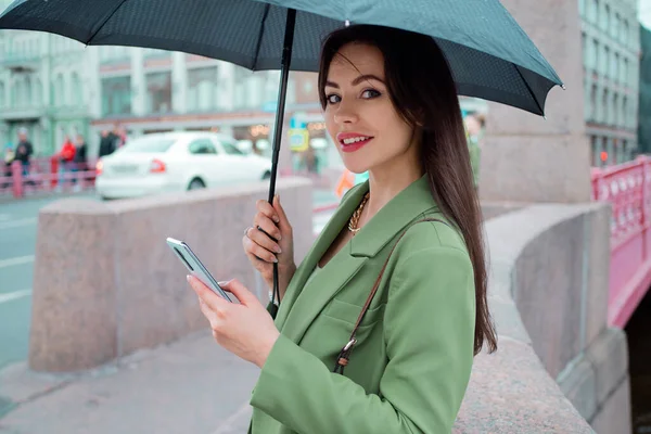 Молодая брюнетка в зеленой куртке на прогулке. Красивая девушка под зонтиком пользуется смартфоном — стоковое фото