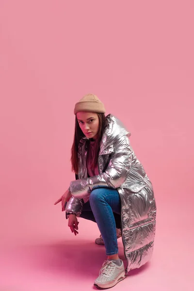 Una chica guay con una chaqueta plateada brillante y un sombrero de punto en cuclillas, — Foto de Stock