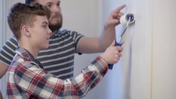 ハンサムな父親は 彼の息子を見せている塗装する方法 幸せな家族の男と十代の少年の家で一緒に修理を行います 父と子絵画壁 白のフラット ローラーを使用して — ストック動画