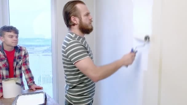 英俊的父亲正在向他的儿子展示如何粉刷墙壁 幸福的家庭 男人和青少年男孩一起在家里做修理 爸爸和孩子画墙在平白色 使用辊 — 图库视频影像