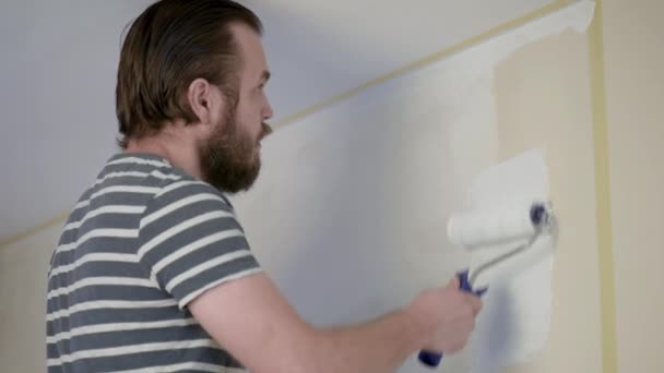ひげを生やした男絵画インテリア壁ペイント ローラーを使用してフラットに ハンサムな若い男 白いペンキで積極的に塗料壁の修理を行います 家の改修や改装のコンセプト — ストック動画
