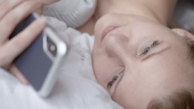 Güzel genç kadın yatıyor yatakta ve akıllı telefon kullanarak. Kadın kısa mesaj yazarak modern cep telefonu sesleri. Çekici kız kaydırma Resimler cep telefonu.