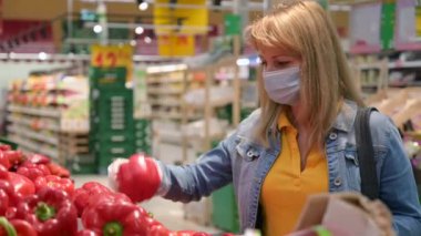 Tıbbi maskeli ve koruyucu eldivenli genç bir kadın kovid-19 Coronavirus salgını sırasında süpermarketten yiyecek seçip alıyor. Yetişkin kız karantina ve kendini izole etme süresince yiyecek stokluyor..