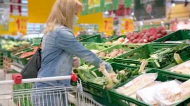 Tıbbi maskeli ve koruyucu eldivenli genç bir kadın kovid-19 Coronavirus salgını sırasında süpermarketten yiyecek seçip alıyor. Yetişkin kız karantina ve kendini izole etme süresince pırasa stokluyor..
