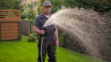 Güzel yeşil çimenleri sulayan bir adam. Erkek çimenlerde yürüyor, su hortumunu tutuyor ve bahçedeki bitkileri suluyor. Bahçıvan ve bahçe fıskiyesi akşam vakti.