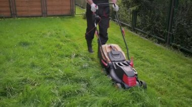 Bahçede çim biçme makinesiyle çim biçen bir adam. İşçi erkek, özel evin bahçesinde yüksek yeşil çim biçiyor. Açık hava mevsimlik ev işleri.