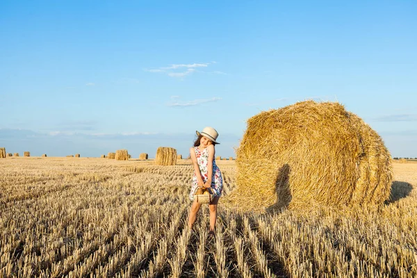 Sevimli kız mutlu bir şekilde yürürken buğday alanında sıcak ve güneşli yaz akşamı hasır şapka giyiyor. Sevimli küçük çocuk çavdar alanında günbatımı ile ekmek sepeti.