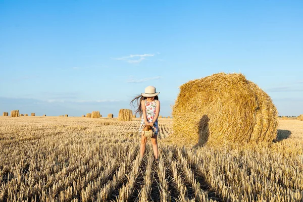Sevimli kız mutlu bir şekilde yürürken buğday alanında sıcak ve güneşli yaz akşamı hasır şapka giyiyor. Sevimli küçük çocuk çavdar alanında günbatımı ile ekmek sepeti.