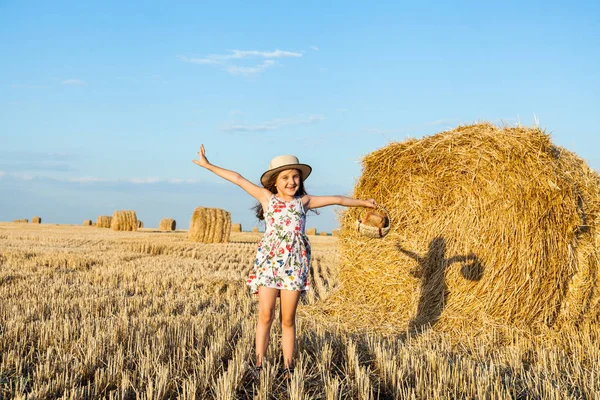 Sevimli kız mutlu bir şekilde yürürken buğday alanında sıcak ve güneşli yaz akşamı hasır şapka giyiyor. Sevimli küçük çocuk çavdar alanında günbatımı ile ekmek sepeti.