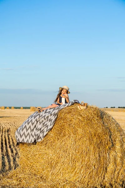 Sevimli kız bir haystack köyü hasır şapka giyiyor süt içip pişmiş mal buğday alanında sıcak ve güneşli yaz akşamı. Sevimli küçük çocuk çavdar alanında günbatımı.