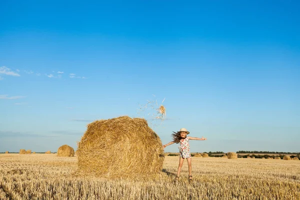 Sevimli kız hasır şapka giyen atlama ve saman buğday alanında kadar sıcak ve güneşli yaz akşamı atma.