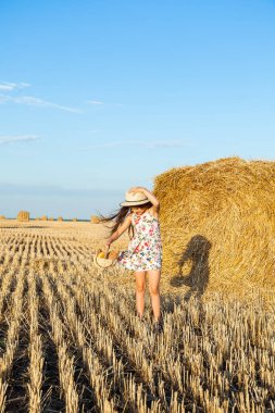 Sevimli kız mutlu bir şekilde yürürken buğday alanında sıcak ve güneşli yaz akşamı hasır şapka giyiyor. Sevimli küçük çocuk çavdar alanında günbatımı ile ekmek sepeti.