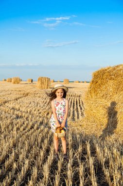 Sevimli kız mutlu bir şekilde yürürken buğday alanında sıcak ve güneşli yaz akşamı hasır şapka giyiyor. Sevimli küçük çocuk çavdar alanında günbatımı ile ekmek sepeti.