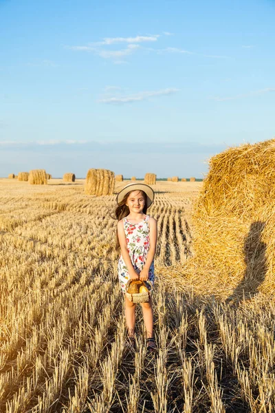 Sevimli kız mutlu bir şekilde yürürken buğday alanında sıcak ve güneşli yaz akşamı hasır şapka giyiyor. Sevimli küçük çocuk çavdar alanında günbatımı ile ekmek sepeti.