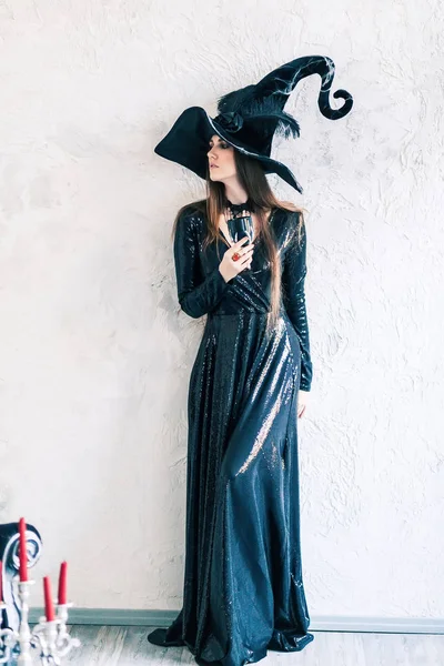Halloween. Portrait of a gorgeous young glamour beautiful woman vampire baroque aristocrat witch in black dress and a hat with a black glass over gray background in the ancient castle.