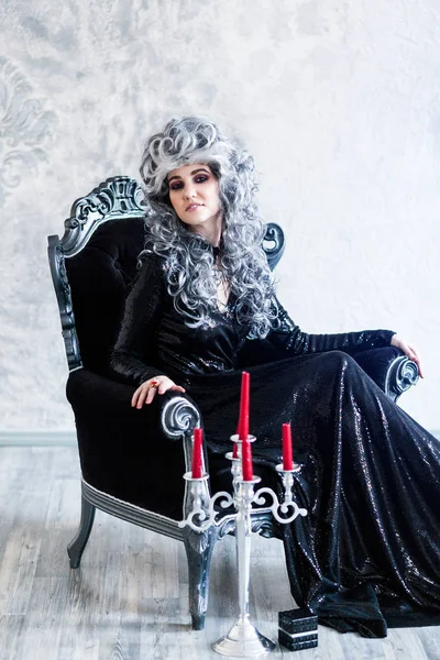 Halloween. Portrait of a gorgeous young glamour beautiful woman vampire baroque aristocrat witch in black dress with gray curly hair with a black glass in the ancient castle.