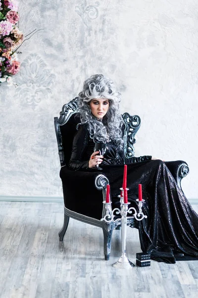 Halloween. Portrait of a gorgeous young glamour beautiful woman vampire baroque aristocrat witch in black dress with gray curly hair with a black glass in the ancient castle.