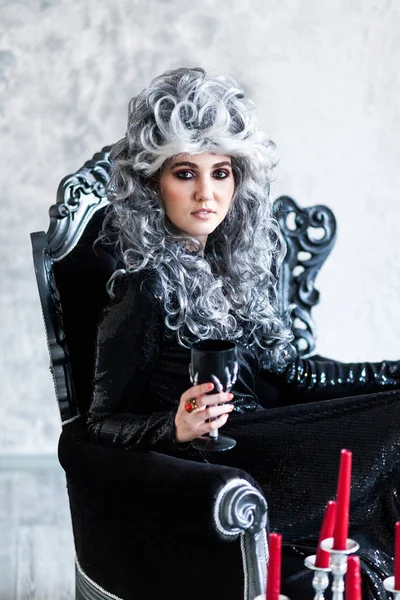 Halloween. Portrait of a gorgeous young glamour beautiful woman vampire baroque aristocrat witch in black dress with gray curly hair with a black glass in the ancient castle.