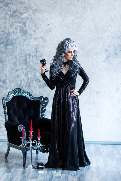 Halloween. Portrait of a gorgeous young glamour beautiful woman vampire baroque aristocrat witch in black dress with gray curly hair with a black glass in the ancient castle.
