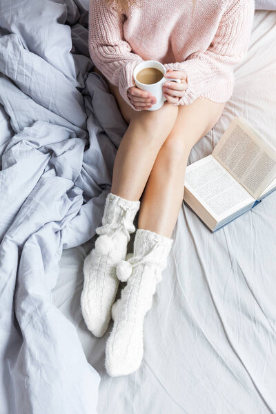 Woman resting keeping legs in warm socks on bed with morning coffee and reading book