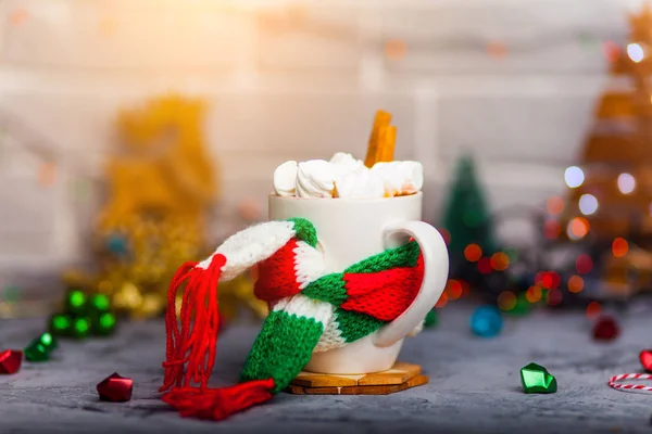 マグカップ ホット コーヒーまたは茶の小型マシュマロとおもちゃでボケ味の美しいクリスマスのイルミネーションで素朴な背景に緑スカーフ白とシナモン — ストック写真