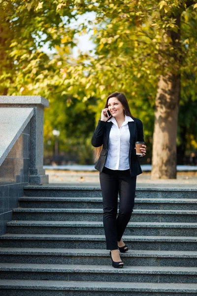 Successful businesswoman with coffee talking on cellphone while walking outdoor. City business woman working.