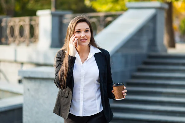 Successful businesswoman with coffee talking on cellphone while walking outdoor. City business woman working.