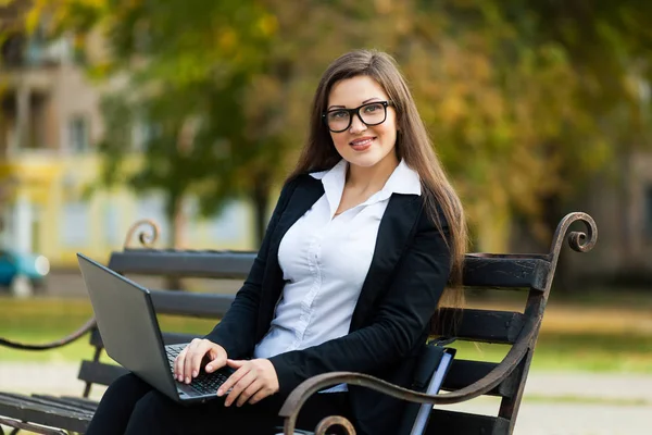 Mulher de negócios sentada no parque em um banco, trabalhando com um laptop — Fotografia de Stock