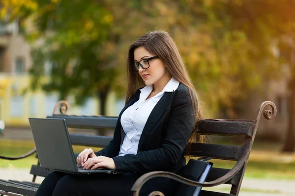 Mulher de negócios sentada no parque em um banco, trabalhando com um laptop — Fotografia de Stock