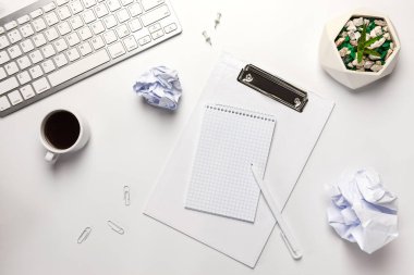 Düz yerde, beyaz ofis masasının üstünde. Çalışma alanı boş klavye, klavye, ofis malzemeleri, kalem, buruşmuş kağıt top, tencerede kaktüs ve beyaz arka planda kahve fincanı..