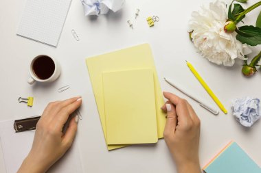 Üst düzey ofis masası ve kadın eli. Klipsli çalışma alanı, sarı defterler, ofis malzemeleri, şakayık çiçekleri ve beyaz arka planda bir fincan kahve..