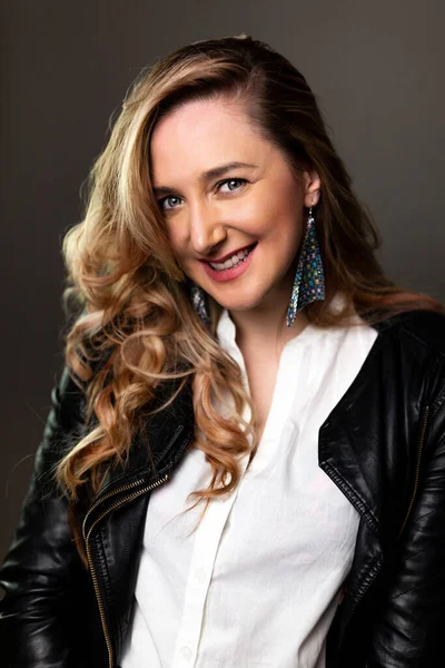 Adult Smiling woman with log curly hair in a white shirt and black leather jacket, studio shot on dark grey wall. Photo with soft focus and film grain