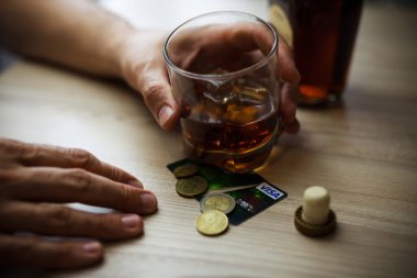 Adam son kuruşuna kadar alkol içiyor. Kırışık alkolik, kahverengi ahşap masada oturuyor, masada bir bardak buzlu konyak, son paralar ve boş kredi kartıyla.